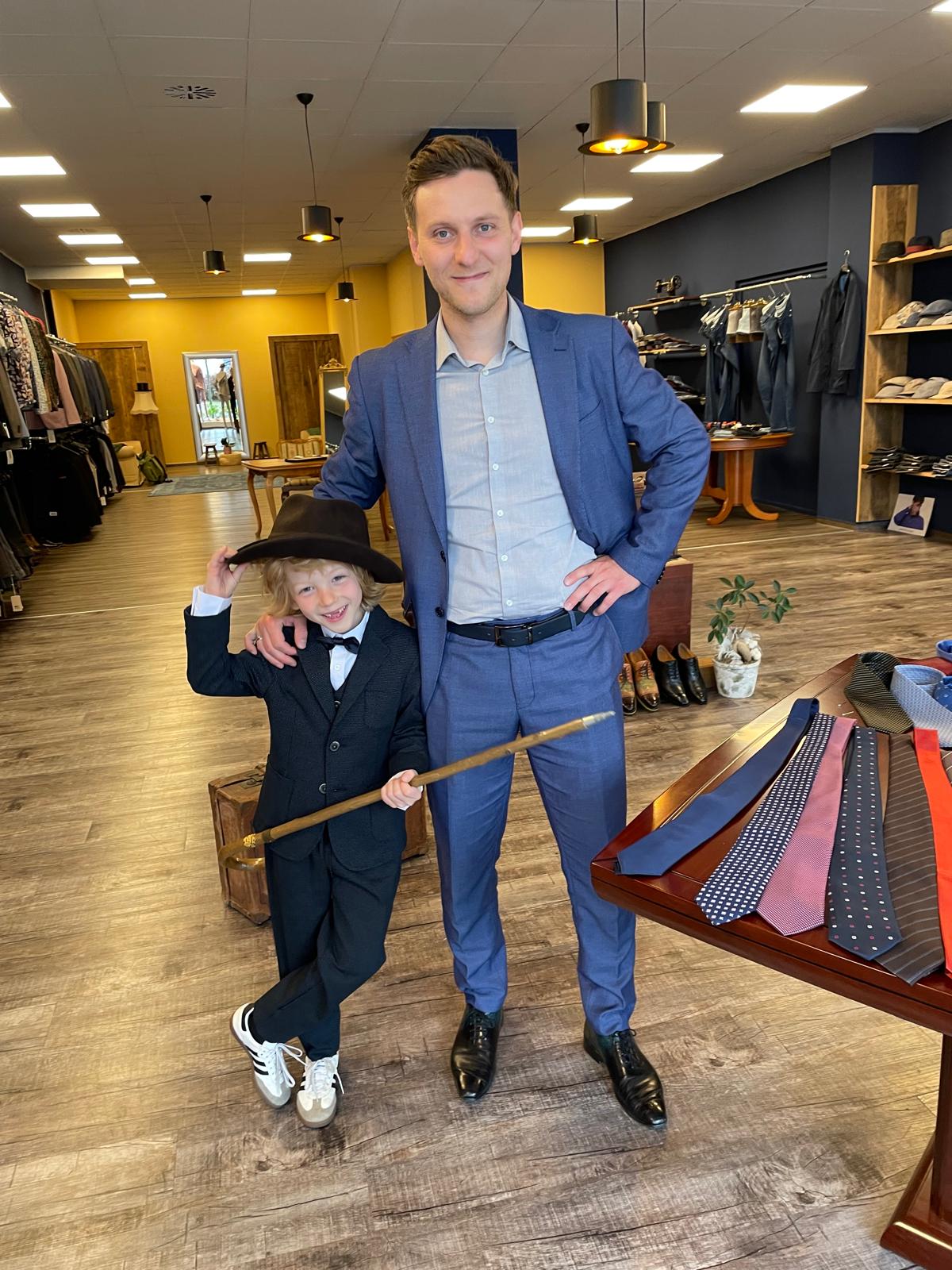 Ein Mann im blauen Anzug steht lächelnd neben einem Kind im schwarzen Anzug mit Hut und Stock