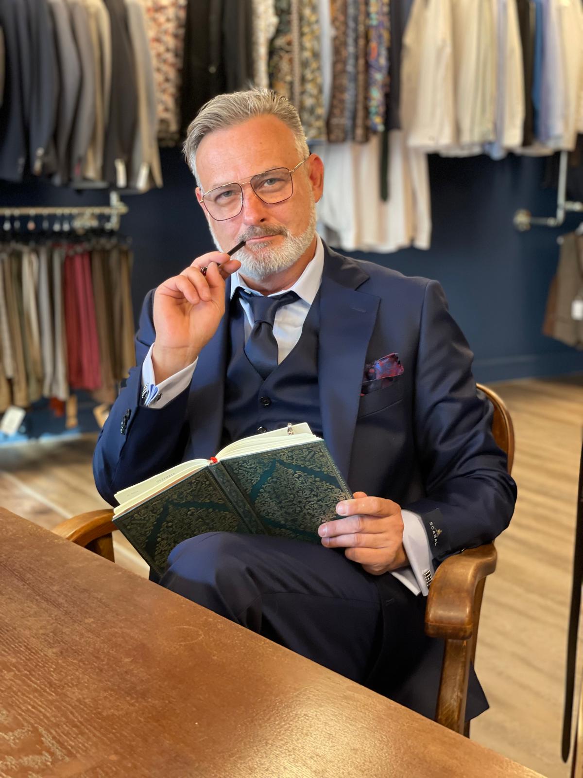 Eleganter älterer Herr mit Brille im blauen Anzug sitzt nachdenklich mit Buch