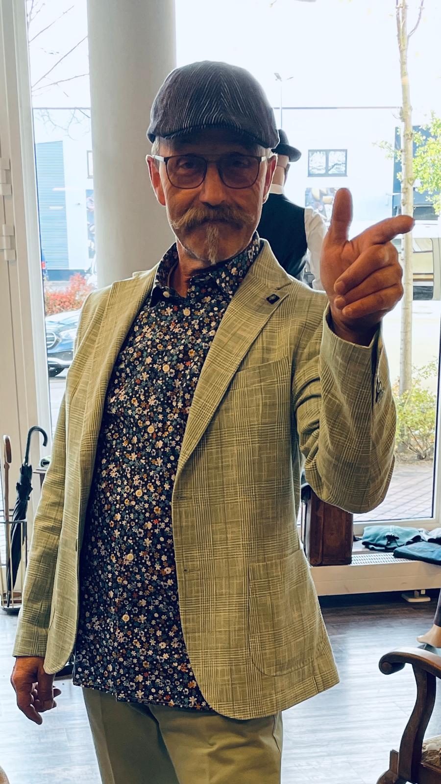 Ein älterer Herr mit grauem Bart und Schnurrbart lächelt verschmitzt und trägt eine helle karierte Jacke sowie eine Schirmmütze