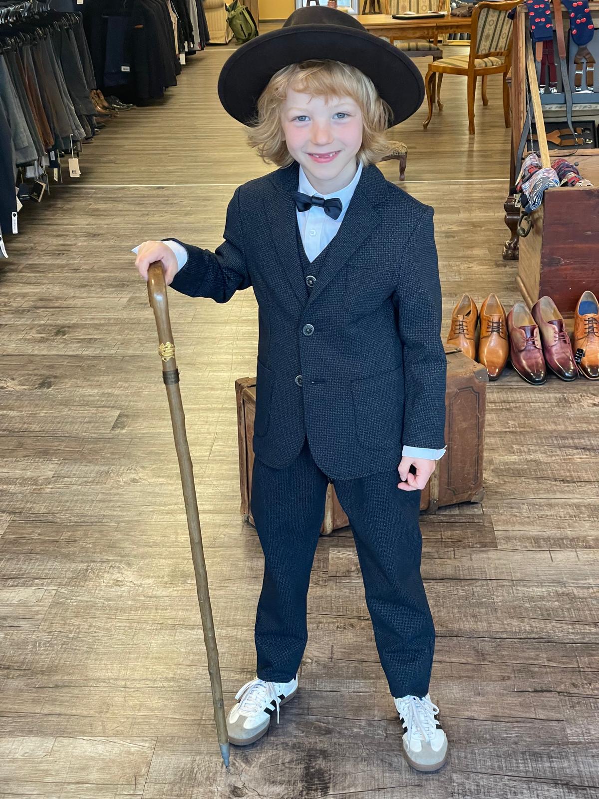 Ein blondes Kind im Anzug und Hut lächelt fröhlich und hält einen Gehstock