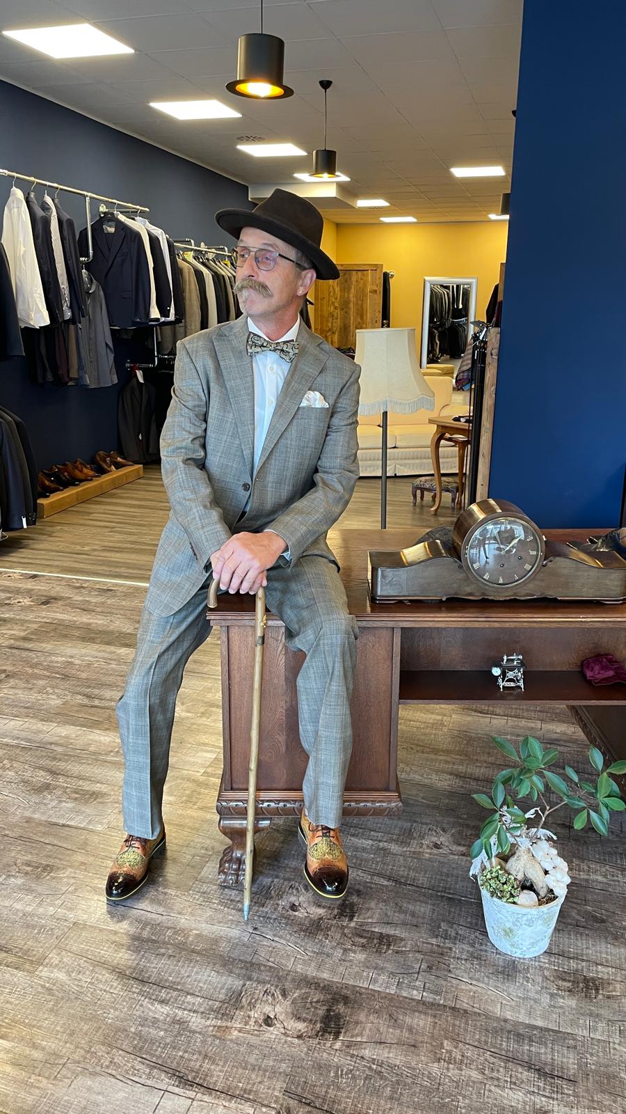 Ein älterer Herr mit grauem Anzug, Fliege und Hut sitzt entspannt mit Stock auf einem Schreibtisch