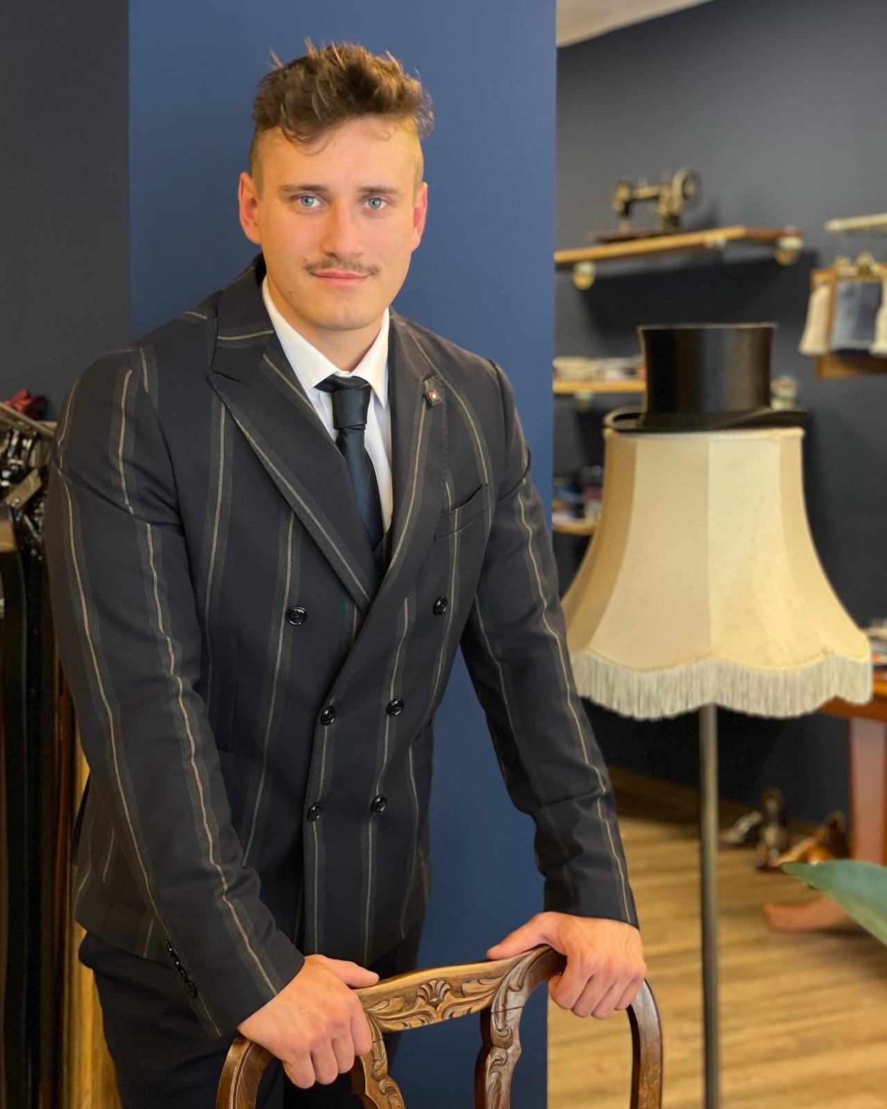 Ein junger Mann mit Schnurrbart trägt einen dunkel gestreiften Anzug und lächelt seriös